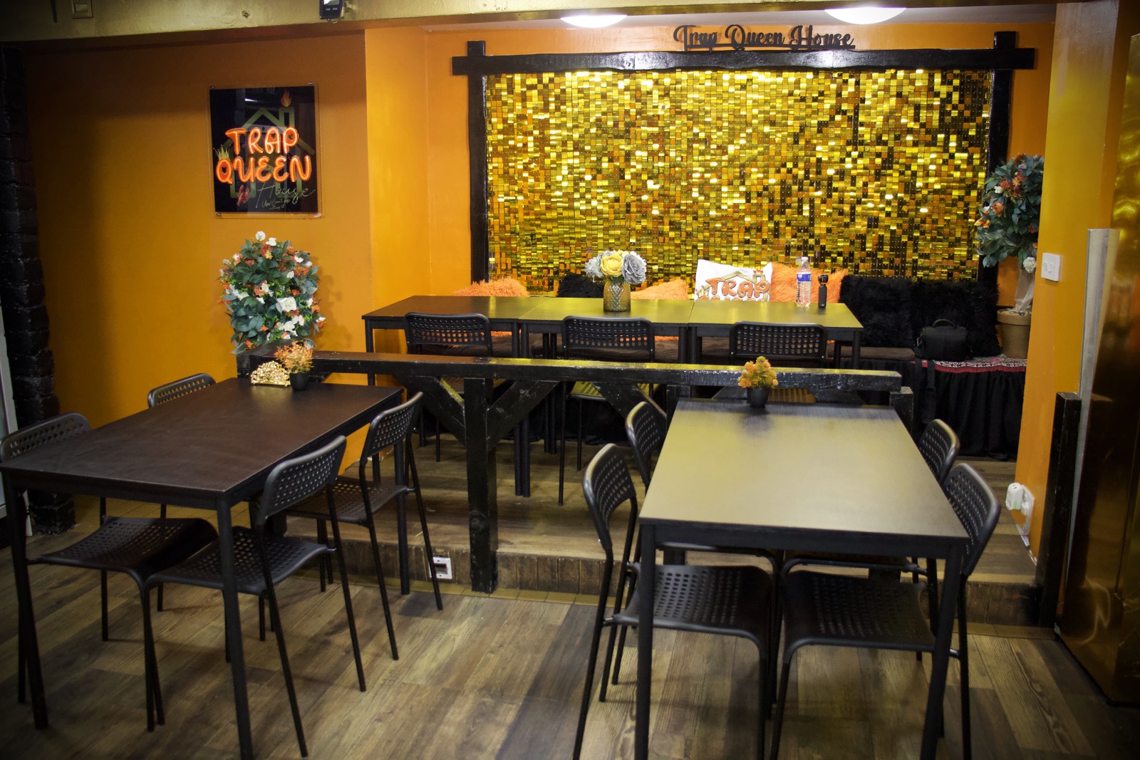 The back room at Trap Queen House — long tables and bench seating under a gold sequin wall, with the neon sign on the orange wall.